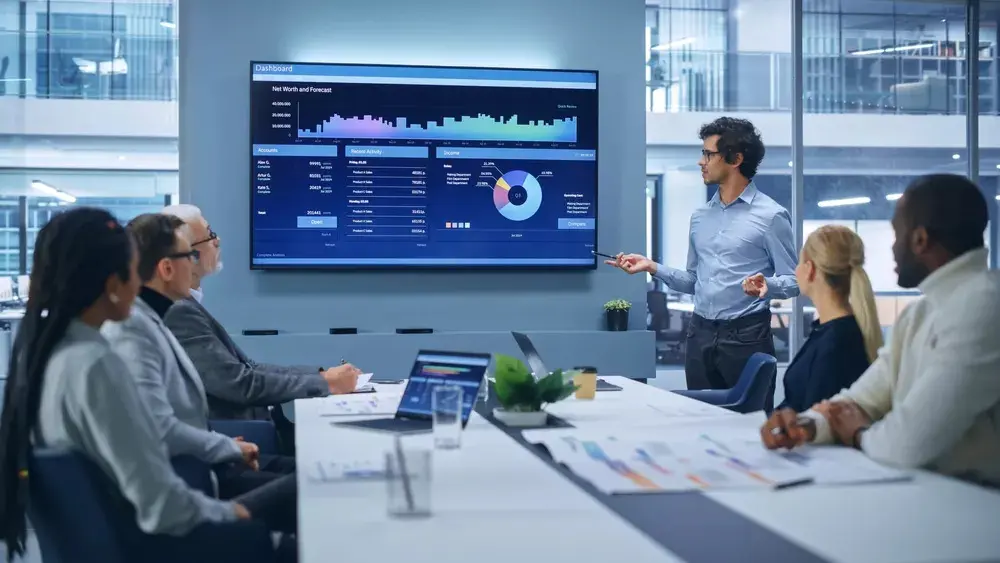 Diverse business team engaged in a presentation with a man pointing to financial data displayed on a large screen in a modern office.