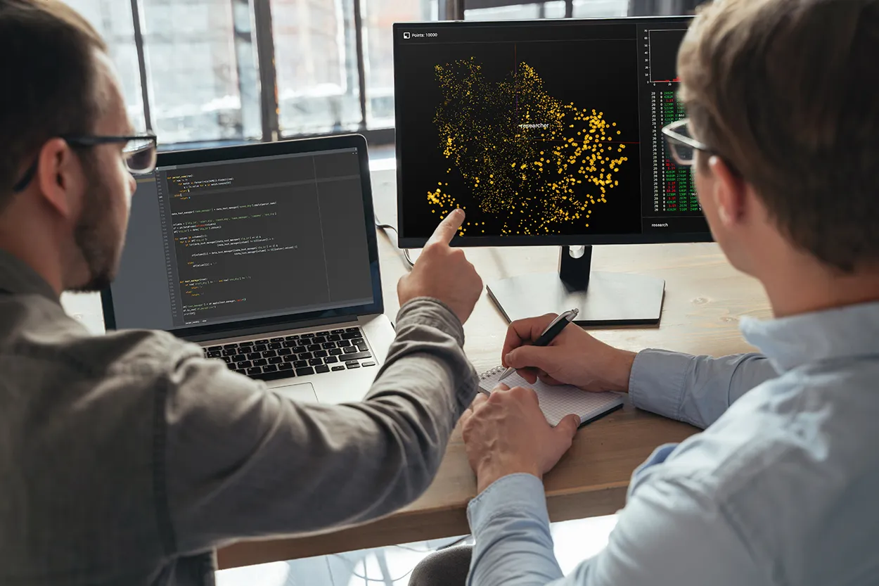 Two colleagues discussing code and data visualization on a laptop and desktop in an office setting