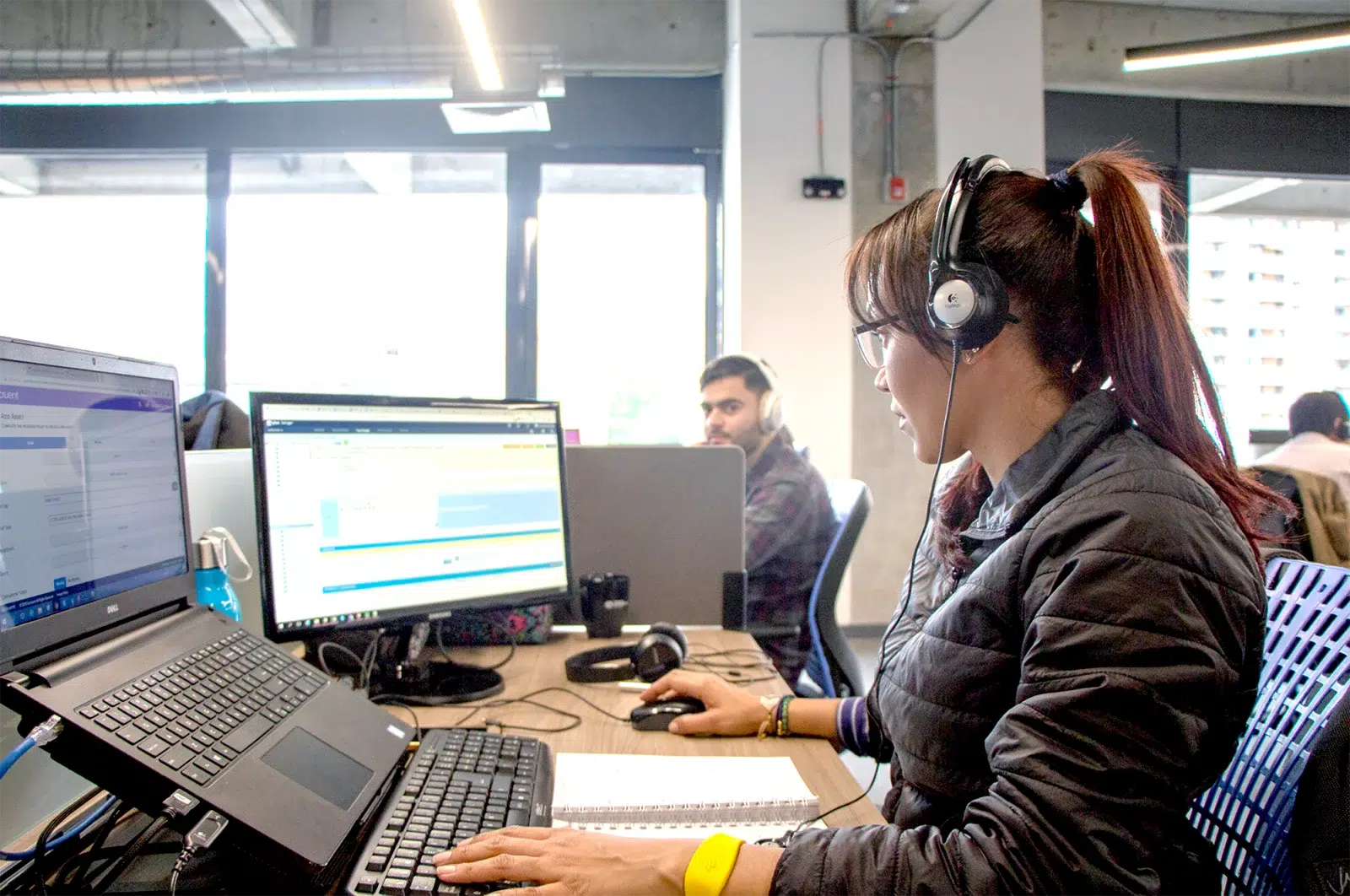 Woman wearing headphones and working at a desk with two monitors, while a colleague sits in the background